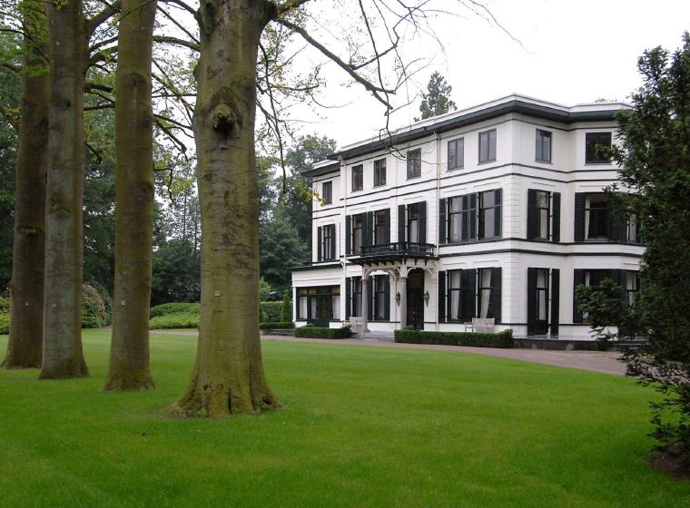 Wit landhuis met groene luiken achter een gazon met hoge bomen