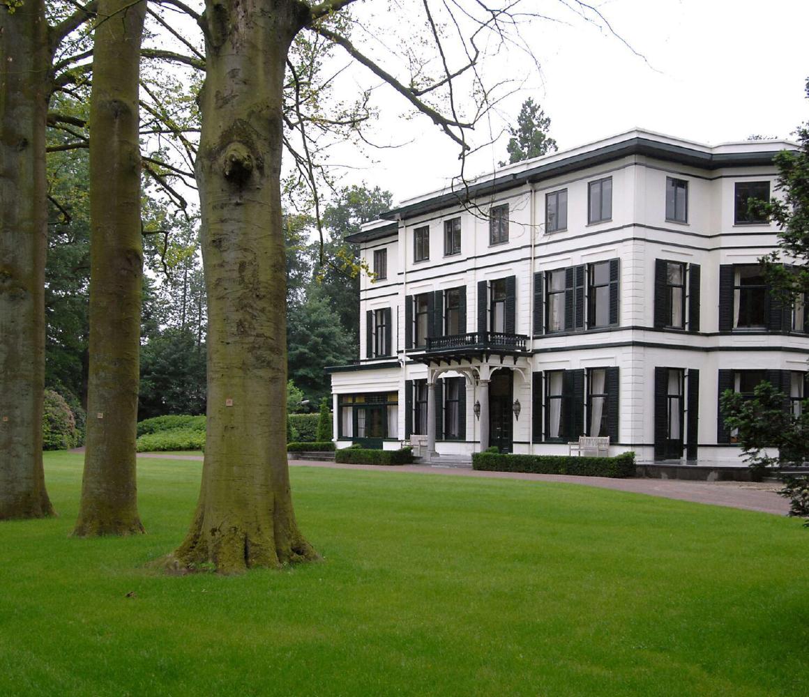 Wit landhuis met groene luiken achter een gazon met hoge bomen