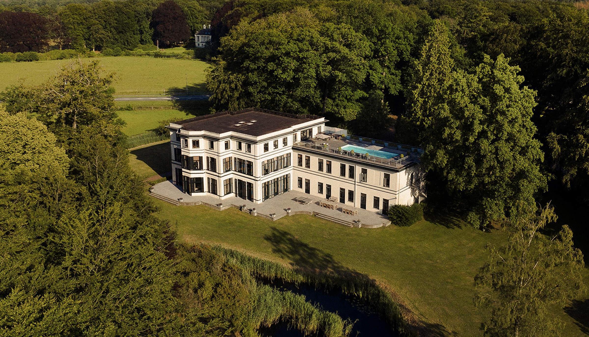 Luchtfoto wit gebouw met pool omgeven door bos en meer